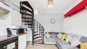A modern kitchen with a built-in oven, a dining area with a red table, and a gray sofa with cushions. - The Mews - 1 Bed Holiday Home - Langland (Swansea)