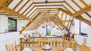 Uma sala de jantar com mesa de madeira posta para quatro pessoas, rodeada por cadeiras de madeira, em um ambiente com teto inclinado e vigas de madeira.