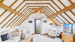 Quarto estilo loft com teto inclinado, vigas de madeira, um sofá, poltronas, uma TV e vista através de uma porta.