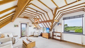 A spacious loft bedroom with a sloped ceiling, a large window offering a view of the countryside, and a cozy seating area with a sofa and armchair. - Hairpin Cottage - 2 Bed Holiday Home - Langland (Swansea)