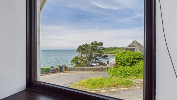 Vista costeira através de uma janela, incluindo uma estrada, uma árvore e um edifício com telhado cónico.