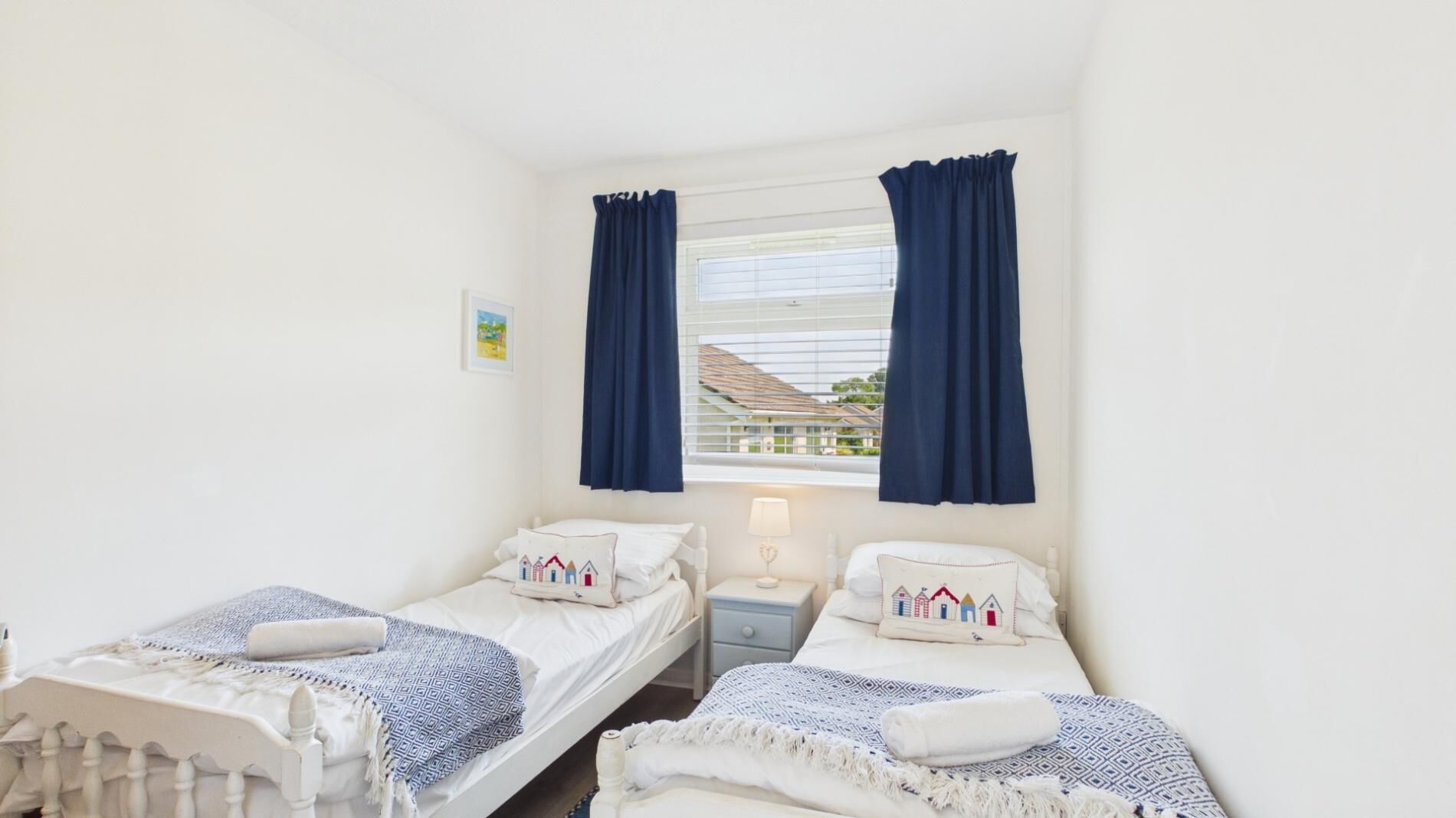 A bedroom with two beds, a window with curtains, a bedside table with a lamp, and a small picture on the wall.