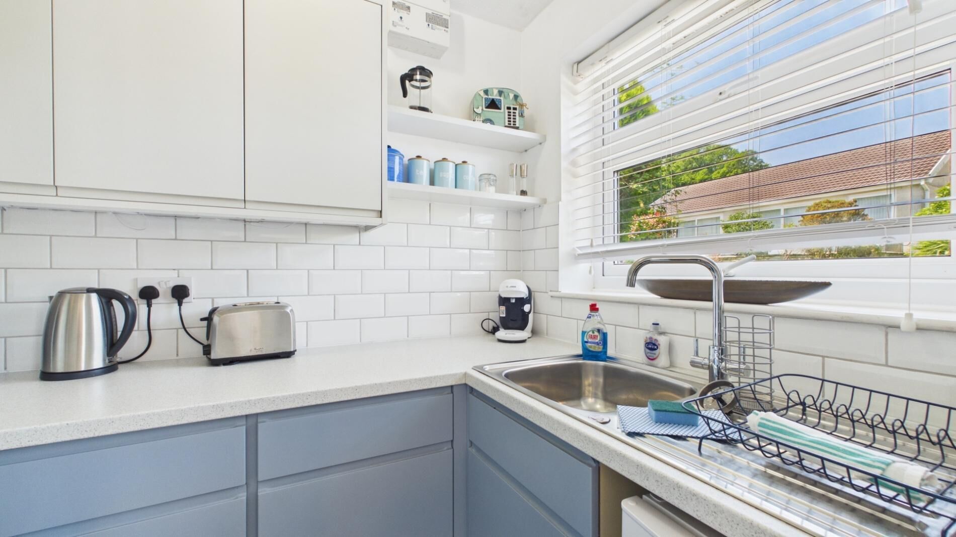 A modern kitchen with white cabinets, a stainless steel kettle, a toaster, a coffee maker, and a window with a view of a house.