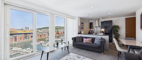 Un apartamento moderno con un ventanal grande, una sala de estar con sofá y mesa de centro, una cocina con armarios negros, y vistas al puerto.