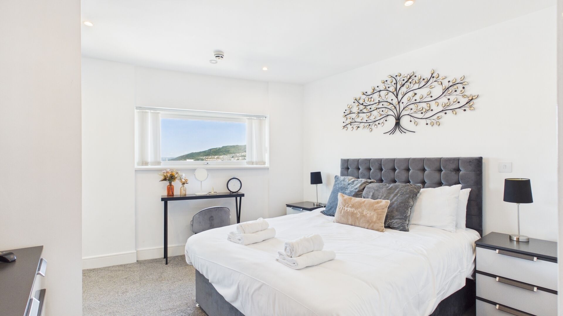 Quarto moderno com cama grande, mesas de cabeceira, escrivaninha e janela com vista.