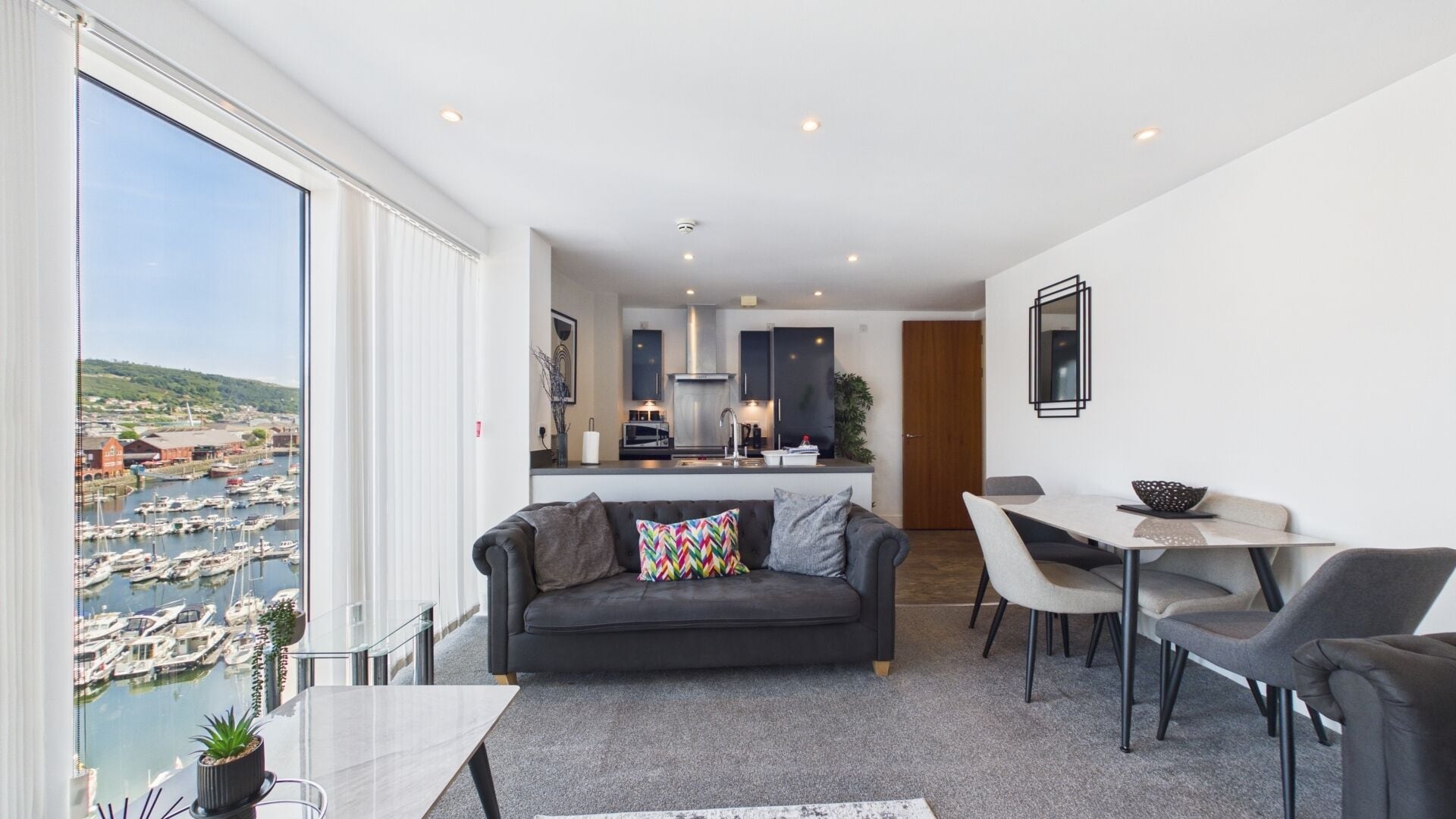 A modern living room with a sofa, dining table, and chairs, a kitchen in the background, and a large window with a marina view.