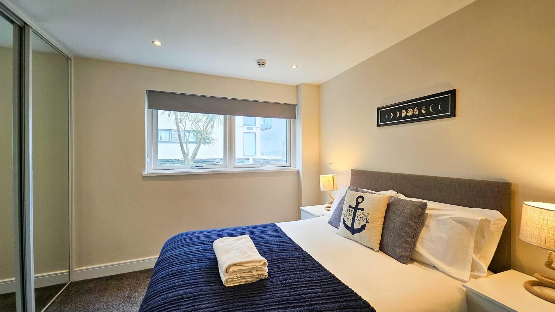 A hotel room with a bed, a window, a wall-mounted artwork, and a nightstand with a lamp.