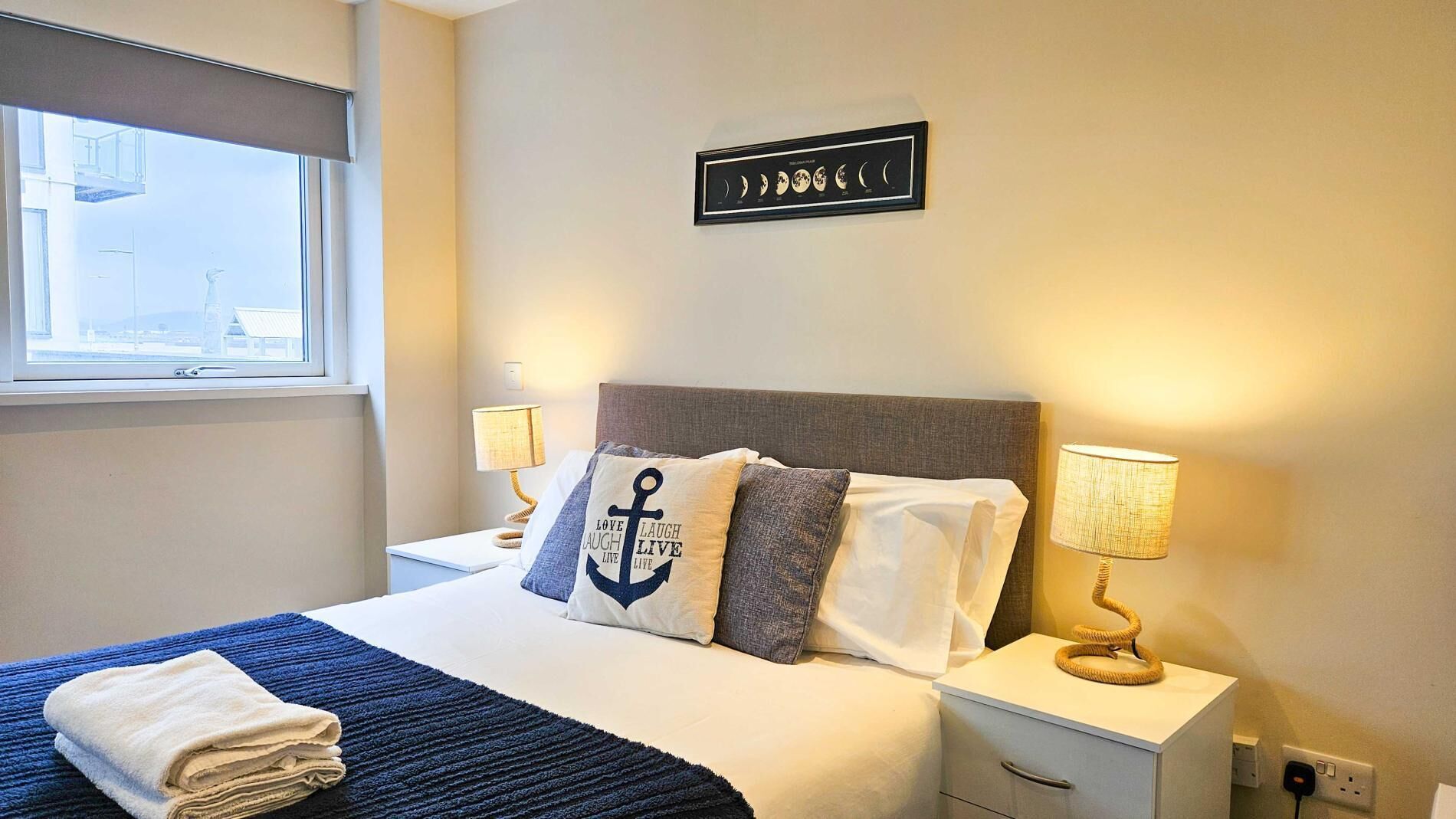 Una cama bien hecha con colcha azul y blanca, dos almohadas con diseño de ancla, un gráfico de fases lunares enmarcado en la pared y una ventana con vista a edificios.
