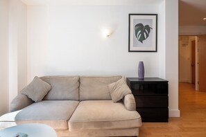 Ein Wohnzimmer mit einem beigefarbenen Sofa, einem schwarzen Beistelltisch mit einer Vase und einer gerahmten Pflanze an der Wand.