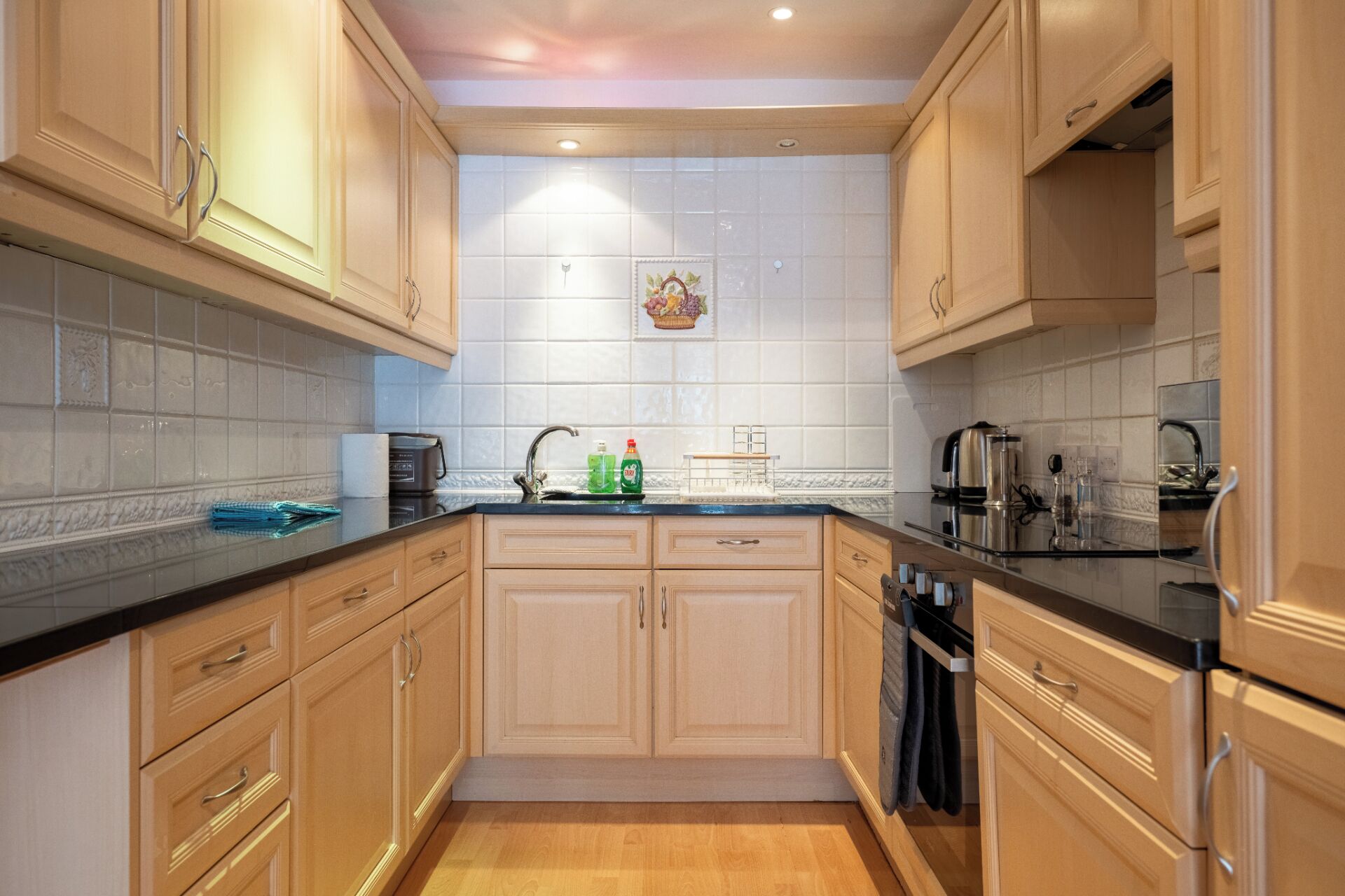 Cocina con alacenas de madera clara, encimera negra y salpicadero de azulejos.
