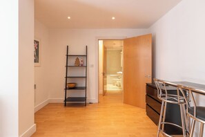 Ein Flur mit Holzboden, der zu einem Badezimmer mit Toilette und Waschbecken führt. Eine Metallreol hat eine Schüssel und eine kleine Zimmerpflanze. Eine Holztür ist teilweise geöffnet, und ein Metallstuhl ist zu sehen.