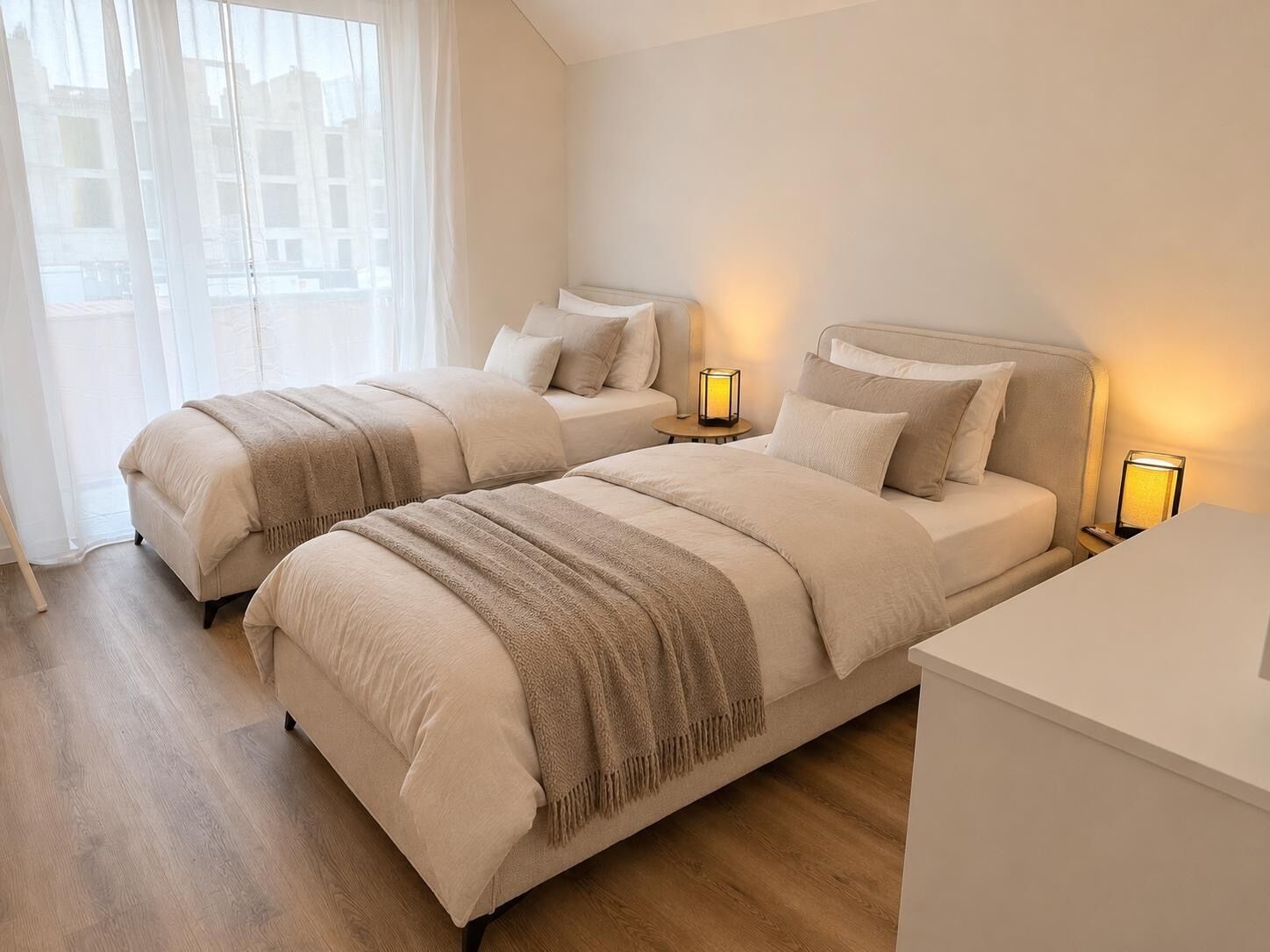 Een slaapkamer met twee bedden, nachtlampjes, een raam met dunne gordijnen en een houten vloer.