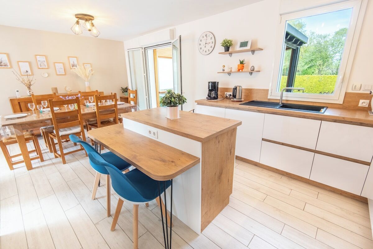 Une cuisine moderne avec un îlot central en bois, un coin repas avec des chaises, une grande fenêtre offrant une belle vue et une horloge au mur.