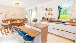 A modern kitchen with a wooden island, a dining area with chairs, a large window with a view, and a clock on the wall. - Family home with a veranda and large garden, 7 min from the beaches by Groom* (Guidel)