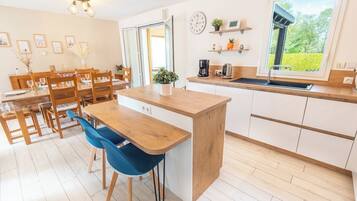 Une cuisine moderne avec un îlot central en bois, un coin repas avec des chaises, une grande fenêtre offrant une belle vue et une horloge au mur.