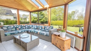 A covered patio area with a sofa set, a glass-top coffee table, and a wooden side table with a potted plant. - Family home with a veranda and large garden, 7 min from the beaches by Groom* (Guidel)