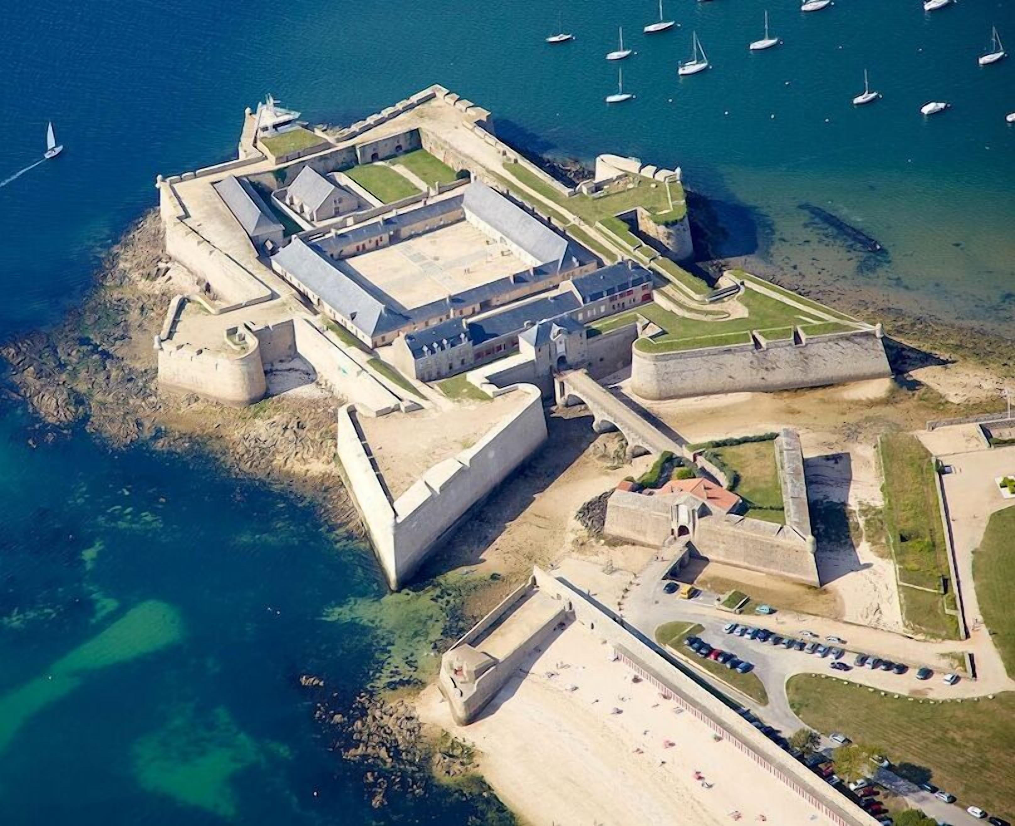 Eine Vogelperspektive einer befestigten Küstenanlage mit mehreren Gebäuden, einem großen Innenhof und einer Marina mit Booten.