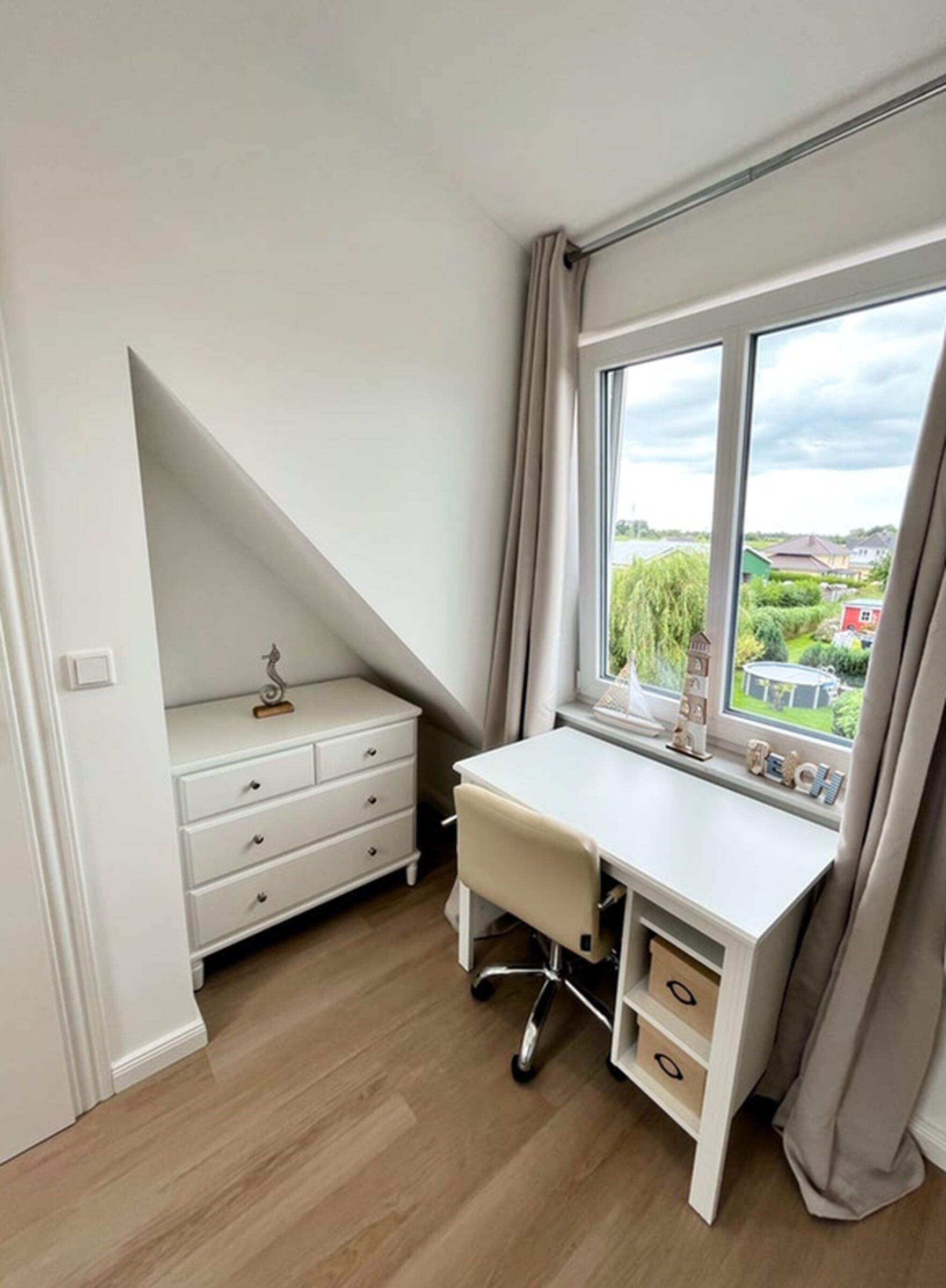 A bedroom with a white desk, a dresser, a window with curtains, and a view of a green landscape.