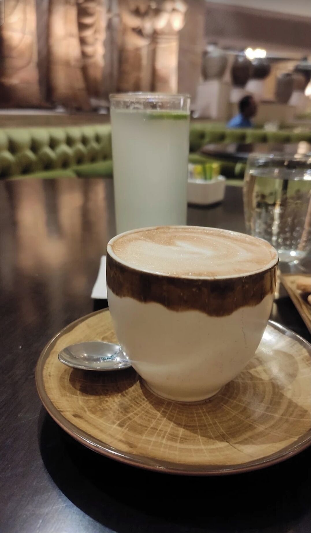 A cup of coffee with a spoon on a wooden coaster, a glass of water, and a green cushioned bench in the background.