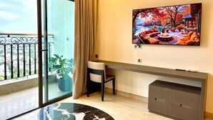 A modern living room with a large TV, a sofa, a desk, and a balcony with a view. - Golden Bay 2 Bedrooms Apartment (Da Nang)