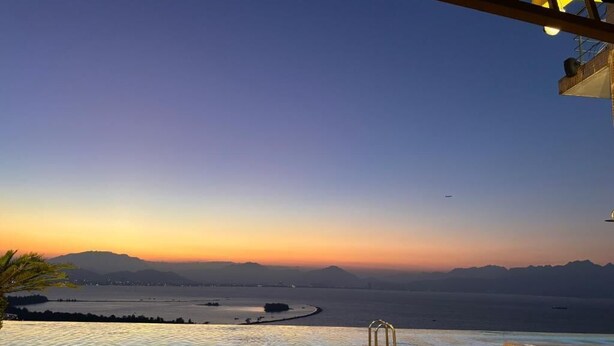 An infinity pool with a view of a sunset over a body of water and distant mountains. - Golden Bay 2 Bedrooms Apartment (Da Nang)