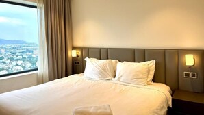 A hotel room with a large bed, two bedside lamps, a view of a cityscape, and a window with curtains. - Golden Bay 2 Bedrooms Apartment (Da Nang)