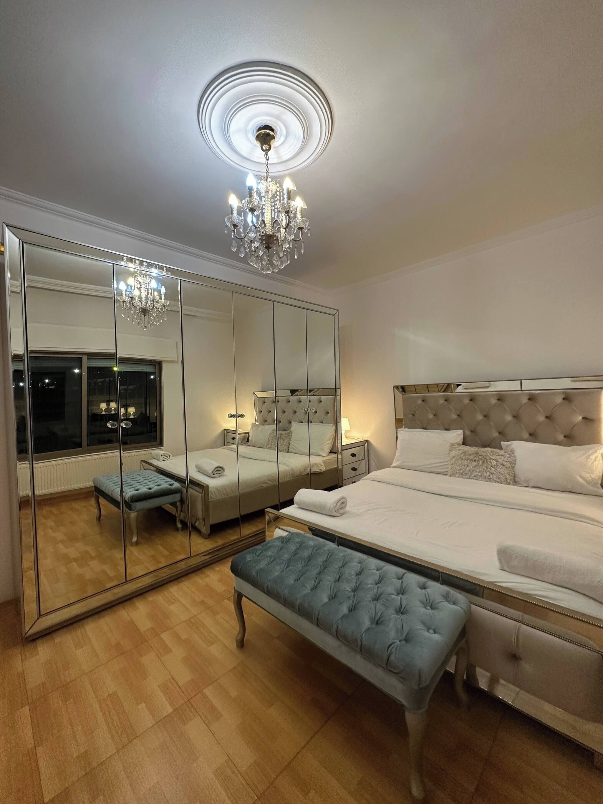 A bedroom with a large mirror, a chandelier, a tufted bed, a bench, and a window.
