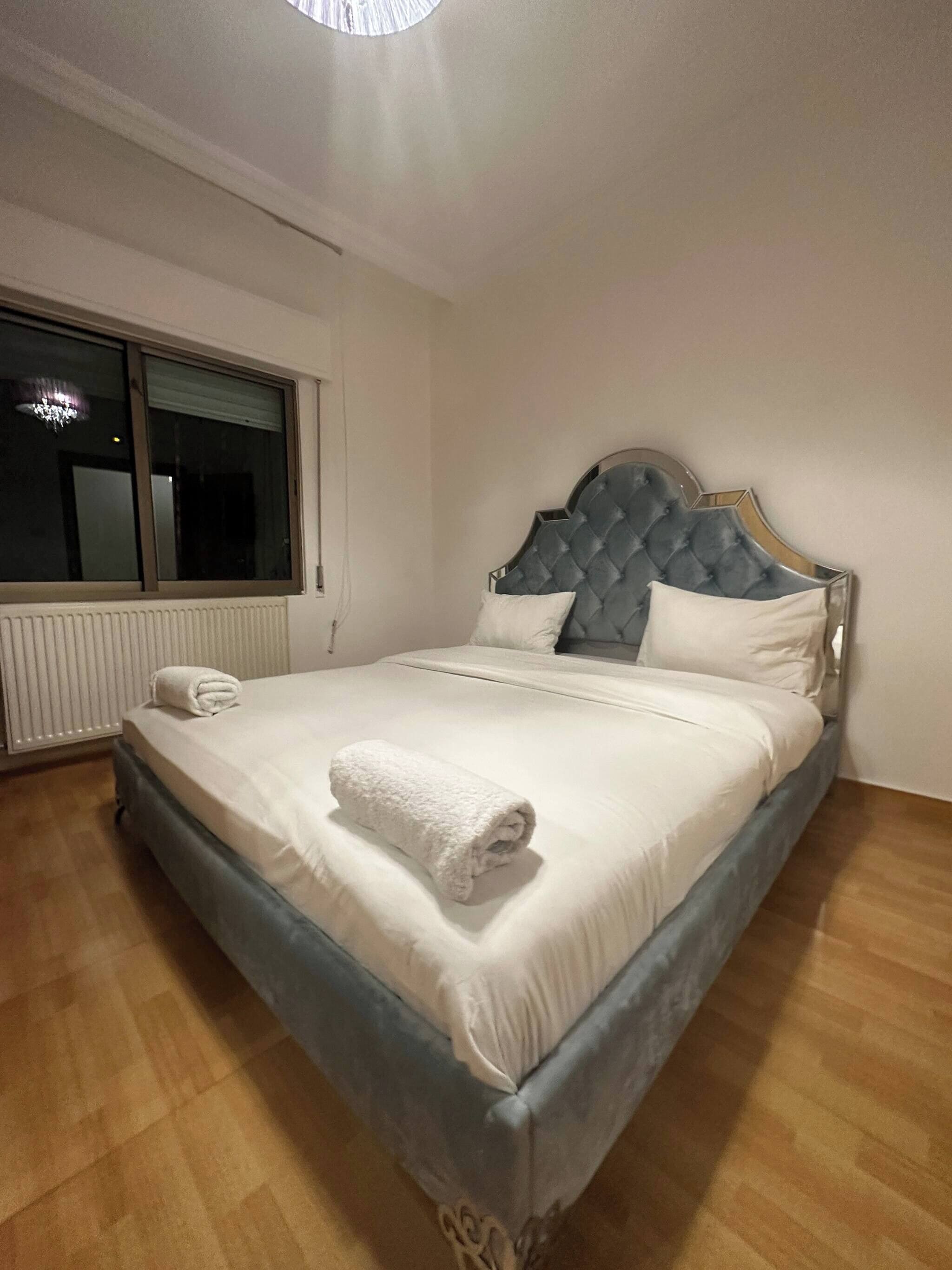Un lit bien fait, avec une serviette blanche roulée posée dessus, une fenêtre avec des stores et un radiateur au mur.