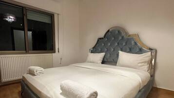 Un lit bien fait, avec une serviette blanche roulée posée dessus, une fenêtre avec des stores et un radiateur au mur.
