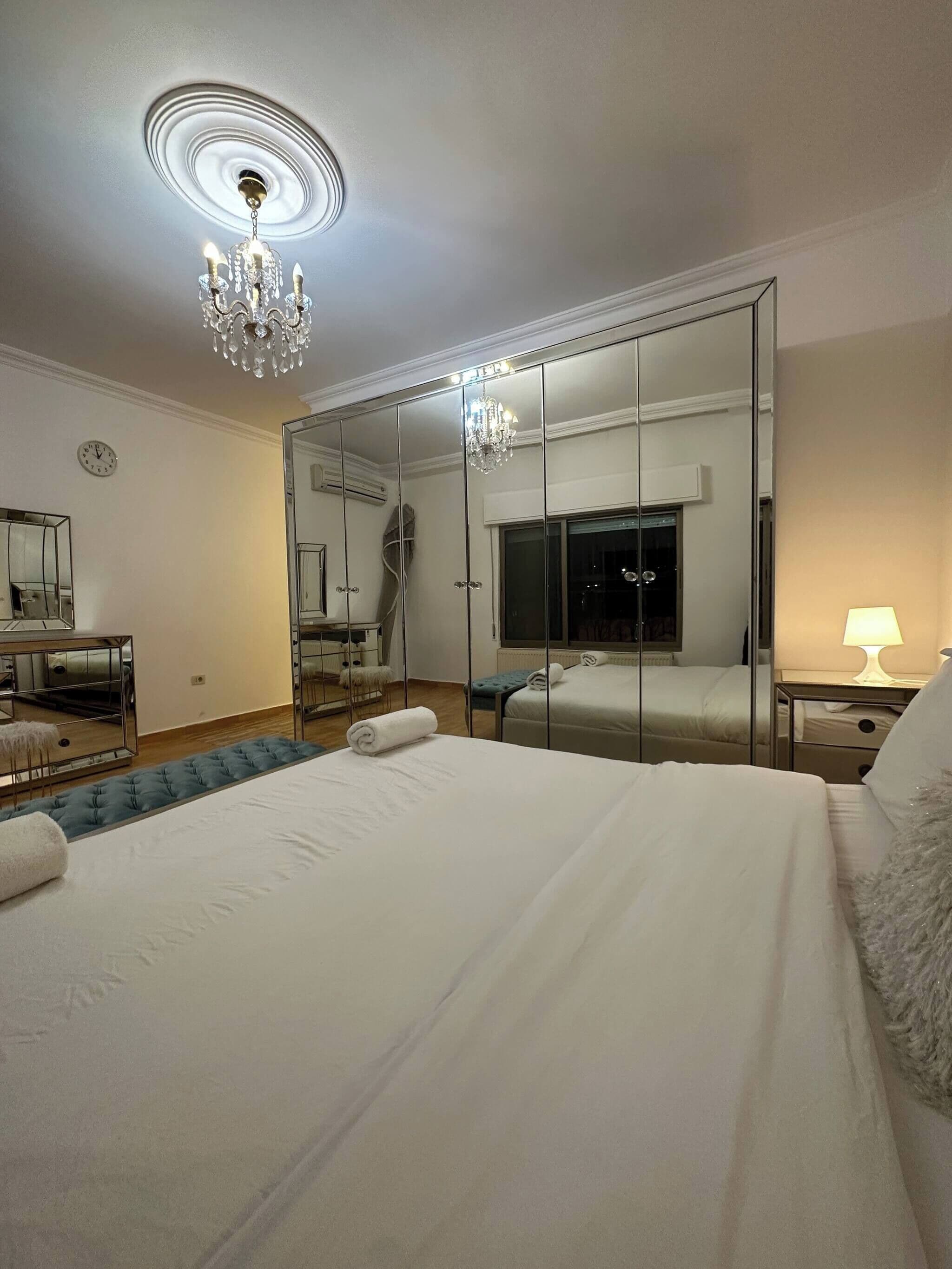 A hotel room with a large bed, a chandelier, a mirror wall, a TV, and a bedside lamp.