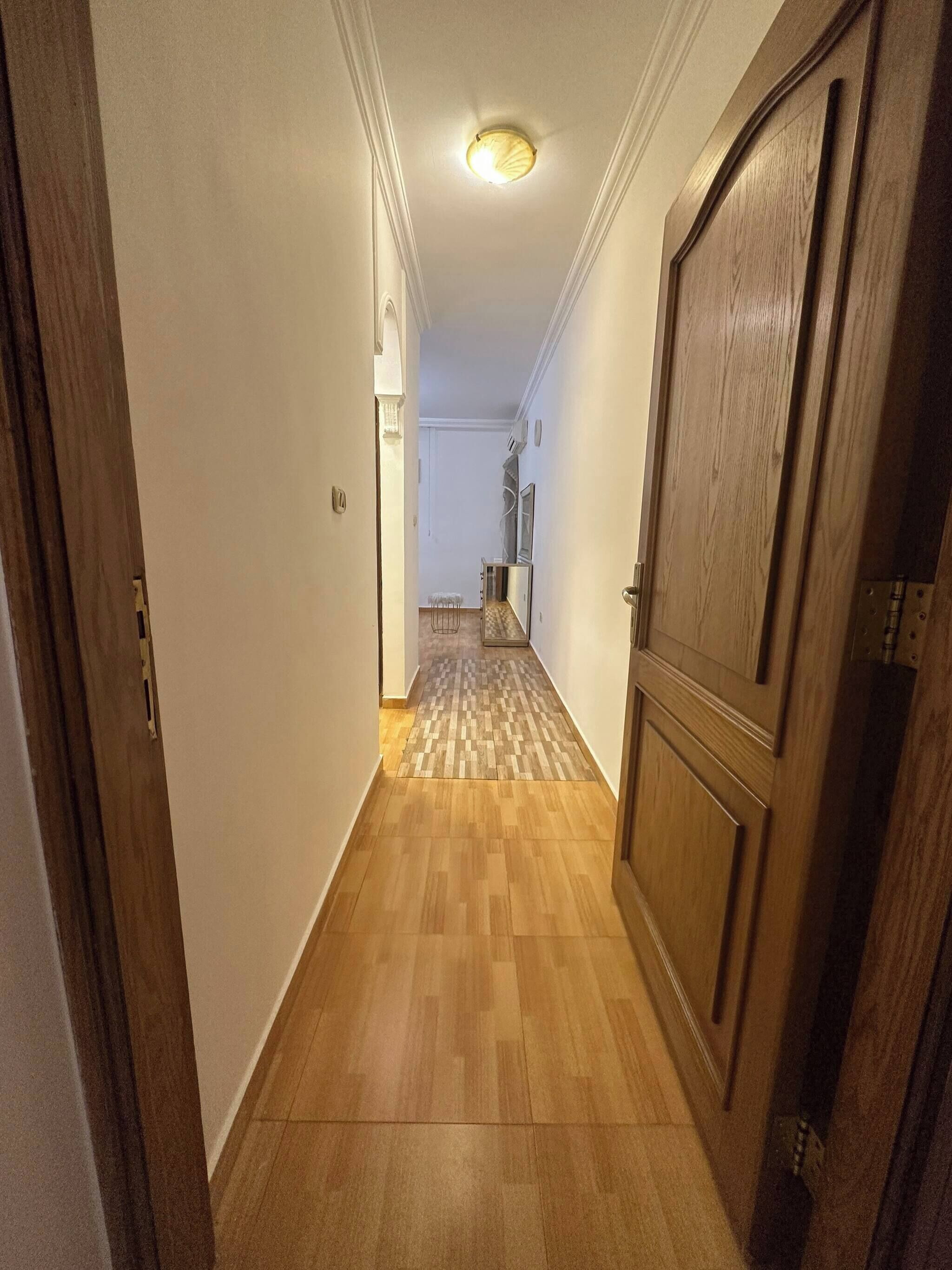 Un pasillo con suelo de madera, una puerta a la derecha y una lámpara en el techo.
