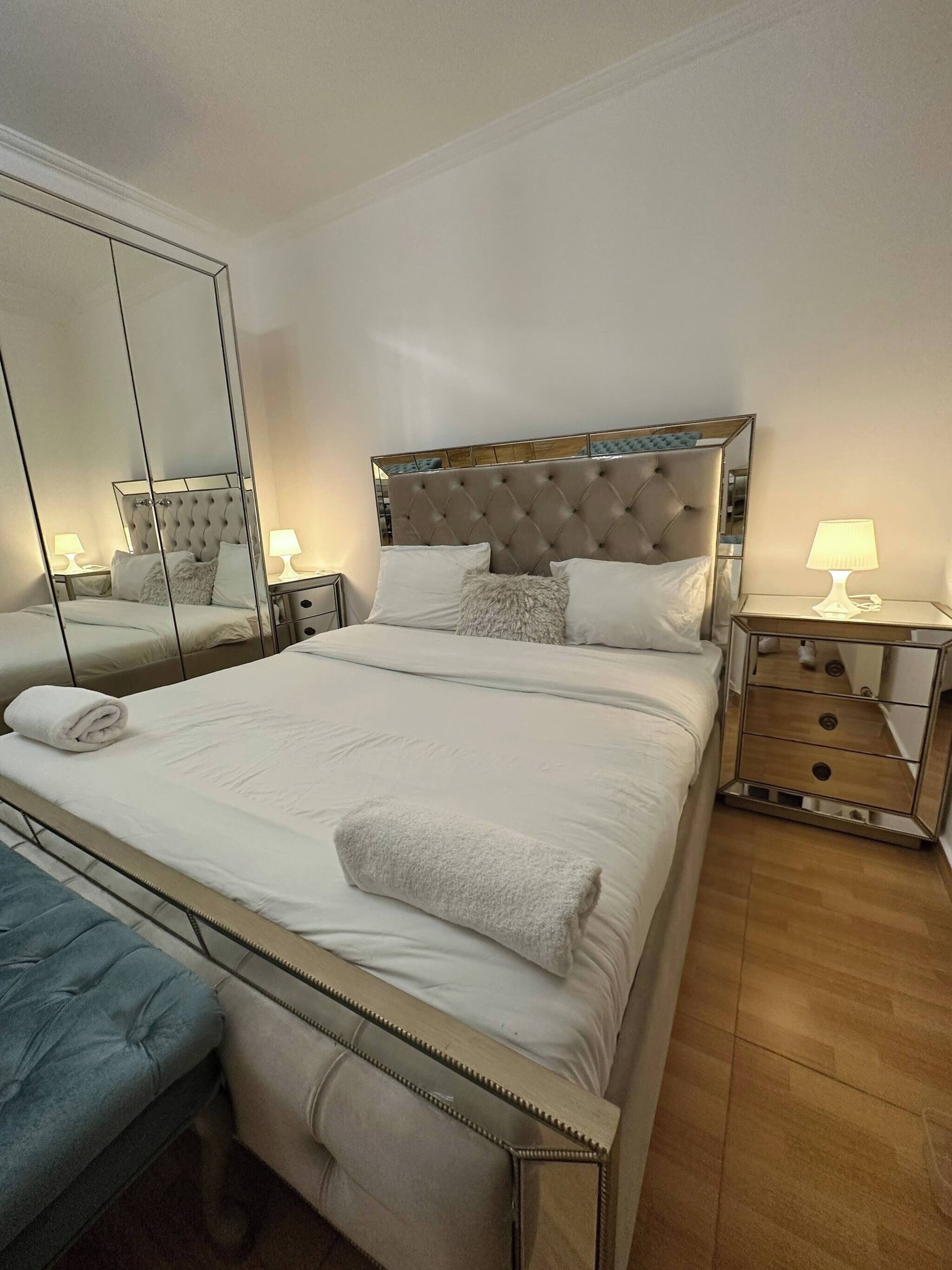 Una cama bien hecha con cabecera capitonada, una mesita de noche con lámpara, un espejo y un banco.