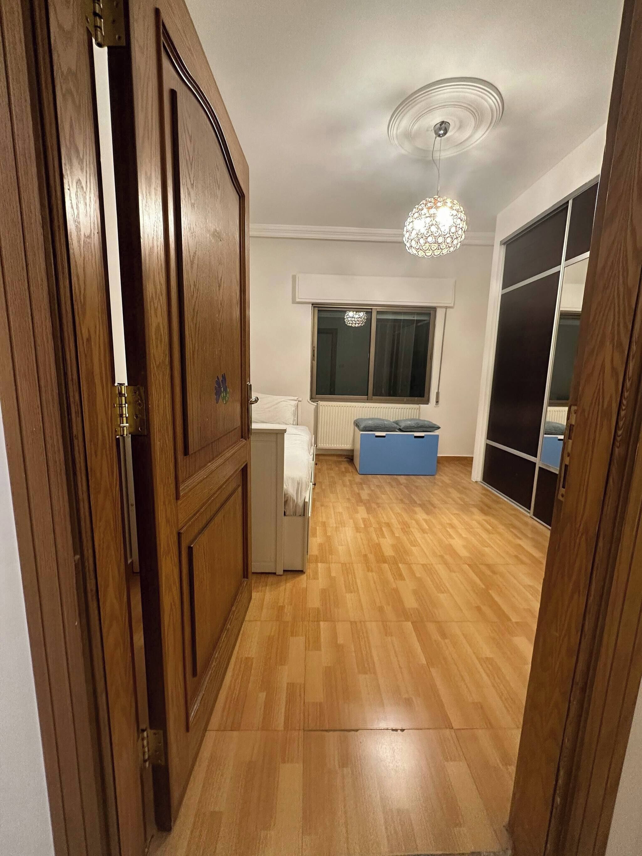A room with a wooden floor, a bed, a dresser, a window with curtains, a ceiling fan, and a chandelier.