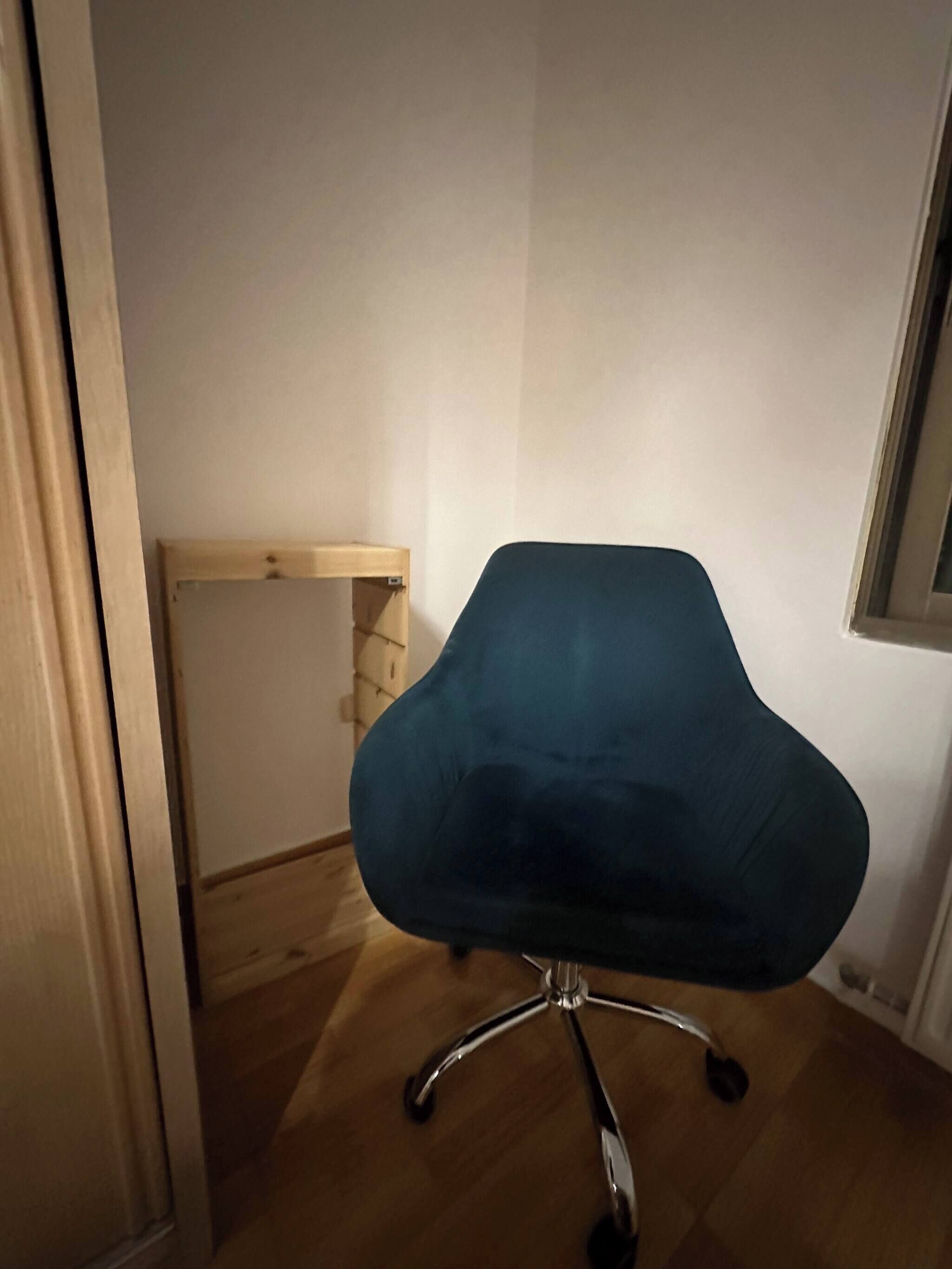 A teal office chair with a five-point base, positioned in a corner of a room with wooden flooring and white walls.
