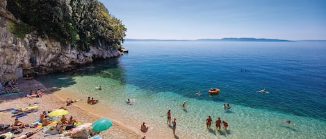 A beach with clear shallow waters, people sunbathing, and a rocky cliff.