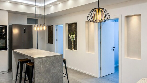 Une cuisine moderne dotée d’un îlot central, de tabourets de bar, de luminaires suspendus et offrant une vue sur une chambre à coucher grâce à une porte ouverte.