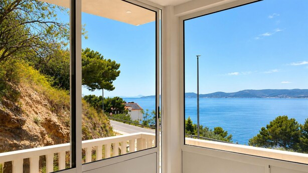 Una stanza con un'ampia finestra che si affaccia su un paesaggio costiero, un balcone con ringhiera e vista sul mare e le colline in lontananza.