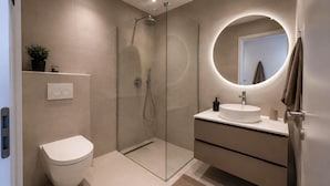 Un baño moderno con ducha de vidrio, inodoro blanco, espejo redondo y lavamanos blanco con encimera color beige.