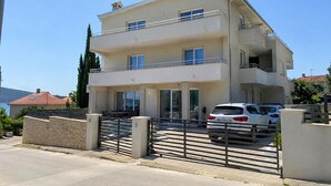 Un edificio residencial de dos plantas con un balcón, un camino de entrada con portón y coches aparcados.