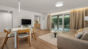 Un soggiorno moderno con una TV a schermo piatto, un'area pranzo con tavolo bianco e sedie in legno, un divano color beige e una porta vetrata che dà su un balcone con tende.