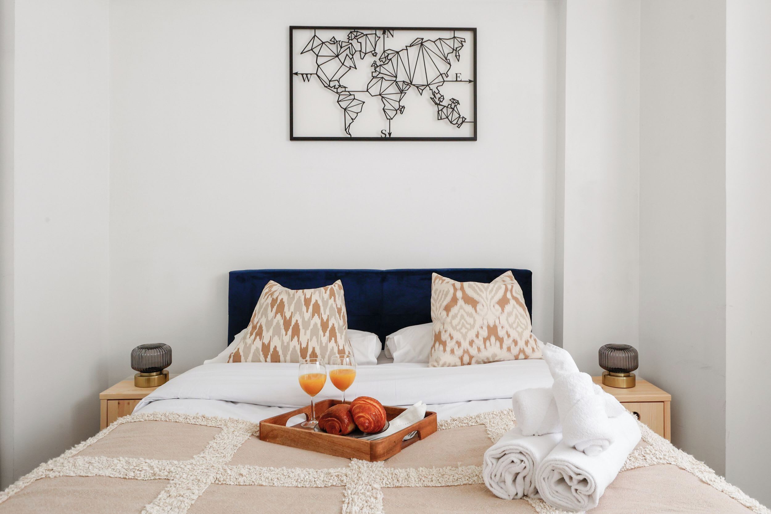 A neatly made bed with a tray of croissants and orange juice, a world map on the wall, and a blue headboard.