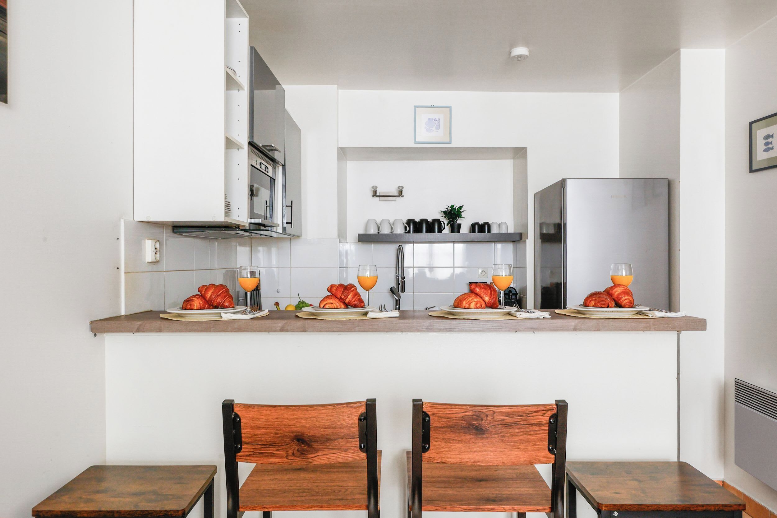 A kitchen with white cabinets, a stainless steel refrigerator, and a breakfast setup with croissants and orange juice.