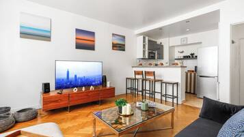 A modern living room with a flat-screen TV, wooden furniture, a glass coffee table, and a kitchen in the background.
