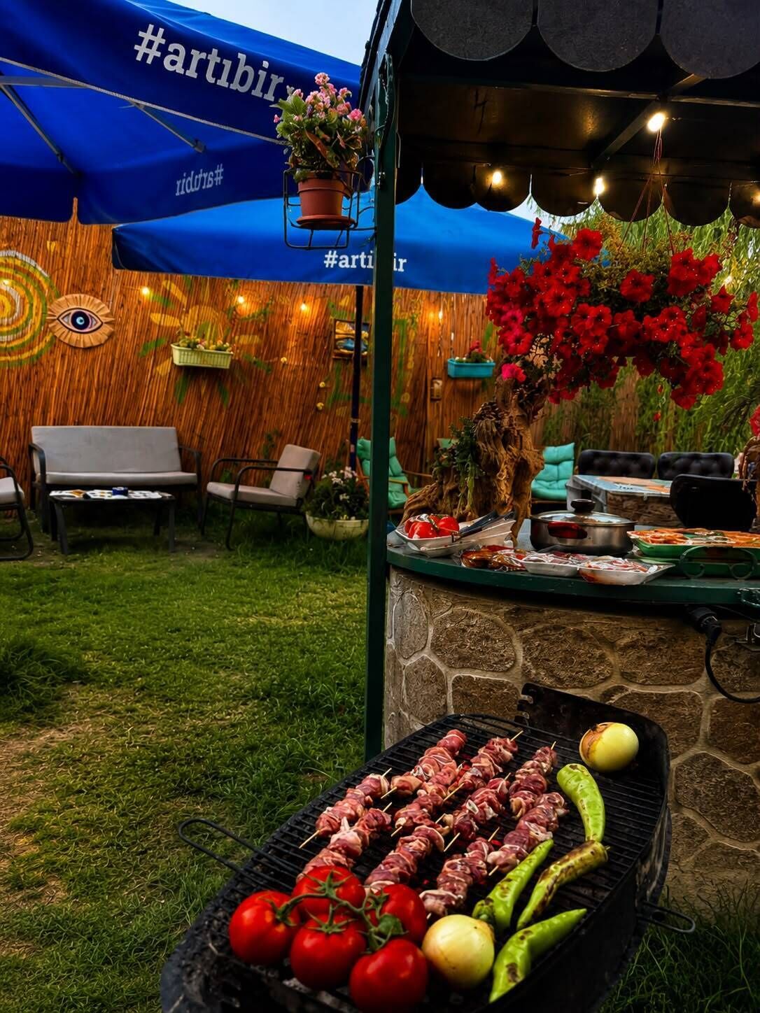 An outdoor barbecue area with a stone counter, a grill with meat skewers and vegetables, and a blue canopy with #artbirr hashtag.