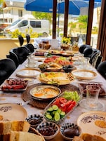 Una mesa dispuesta para una comida con varios platos, incluyendo pan, verduras y carnes, en un espacio exterior para comer con vistas a edificios y una furgoneta al fondo.