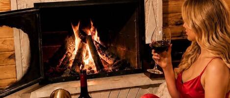 A woman in a red dress sitting by a fireplace with a bottle of wine, fruit, and a decorative wall.