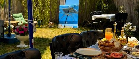 An outdoor dining area with a table set for a meal, surrounded by blue umbrellas, and a view of a garden.