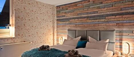 A modern bedroom with a brick-patterned wall, a wooden panel accent wall, a bed with a grey headboard, and a bedside table with a lamp.