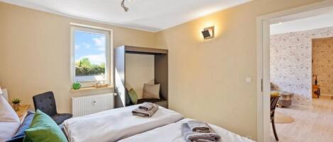 A bedroom with two beds, a window, a radiator, a mirror, and a door leading to a bathroom.
