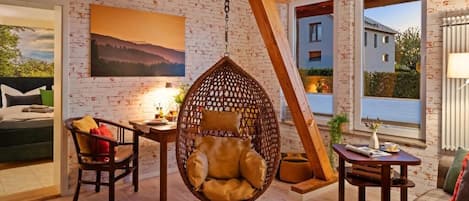 Une pièce avec un fauteuil suspendu, une table avec une lampe, un canapé et un tableau au mur.